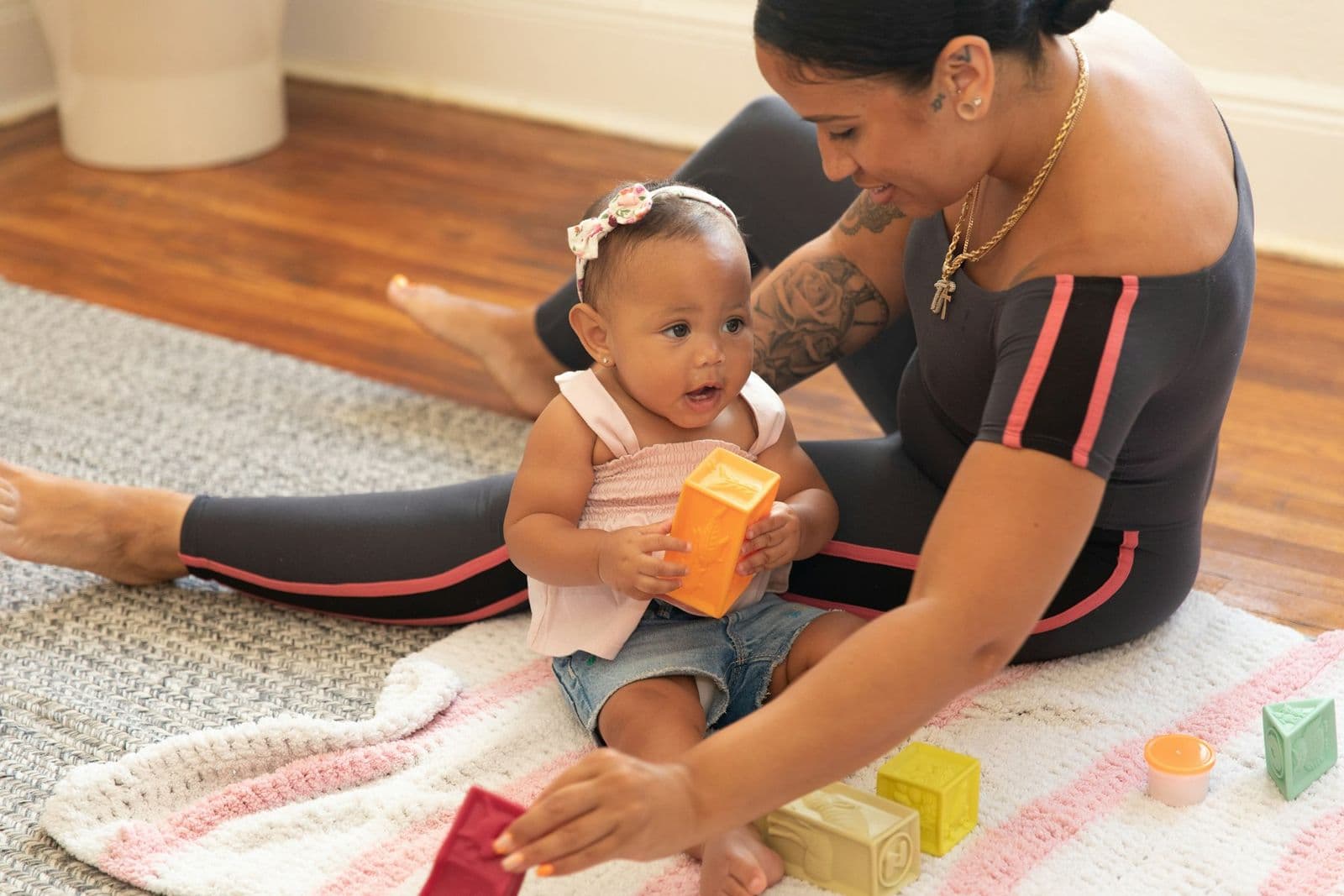 A mother in workout clothes sitting on a rug at home while her baby plays with blocks