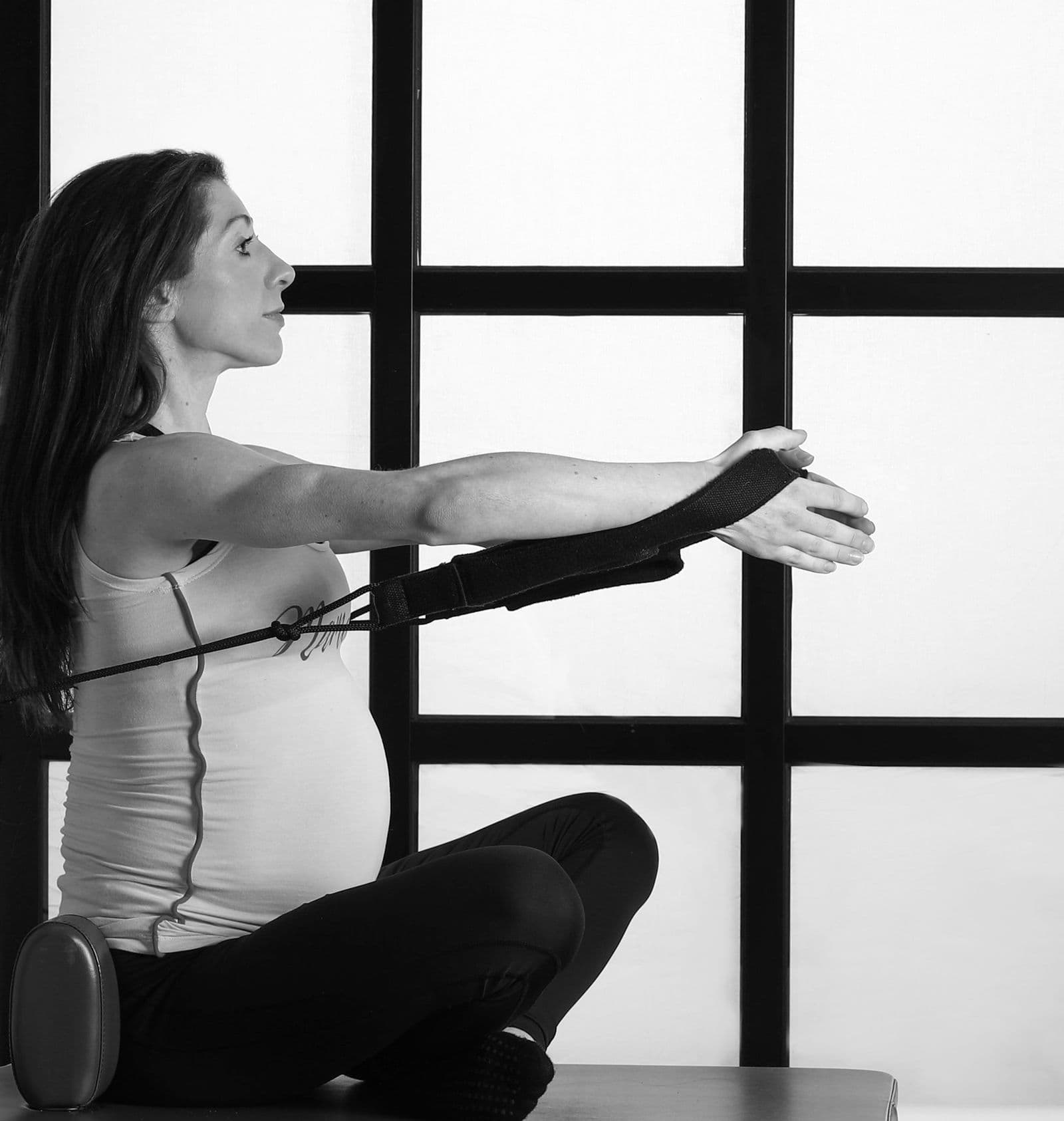 A pregnant woman sitting on a reformer, working her arms with the straps